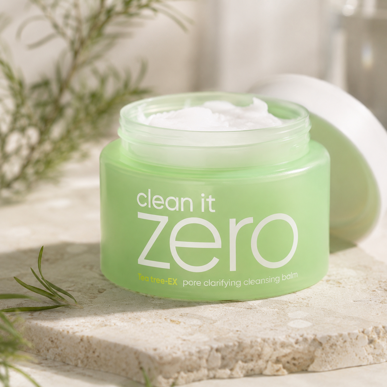 Banila Co Clean It Zero Tea Tree-EX Pore Clarifying Cleansing Balm shown open with its white balm texture on warm travertine in soft natural light.