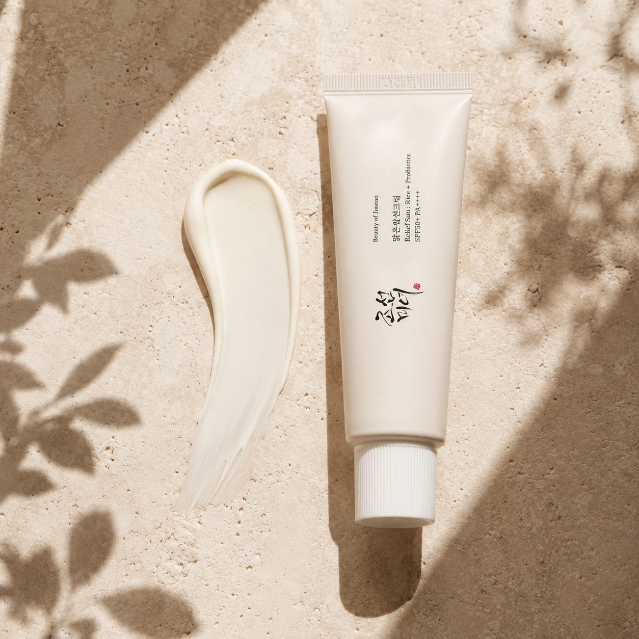 Beauty of Joseon Relief Sun Rice + Probiotics SPF50+ PA++++ 50ml beside a smooth white sunscreen cream texture on warm travertine, showing its lightweight creamy finish in soft natural light.