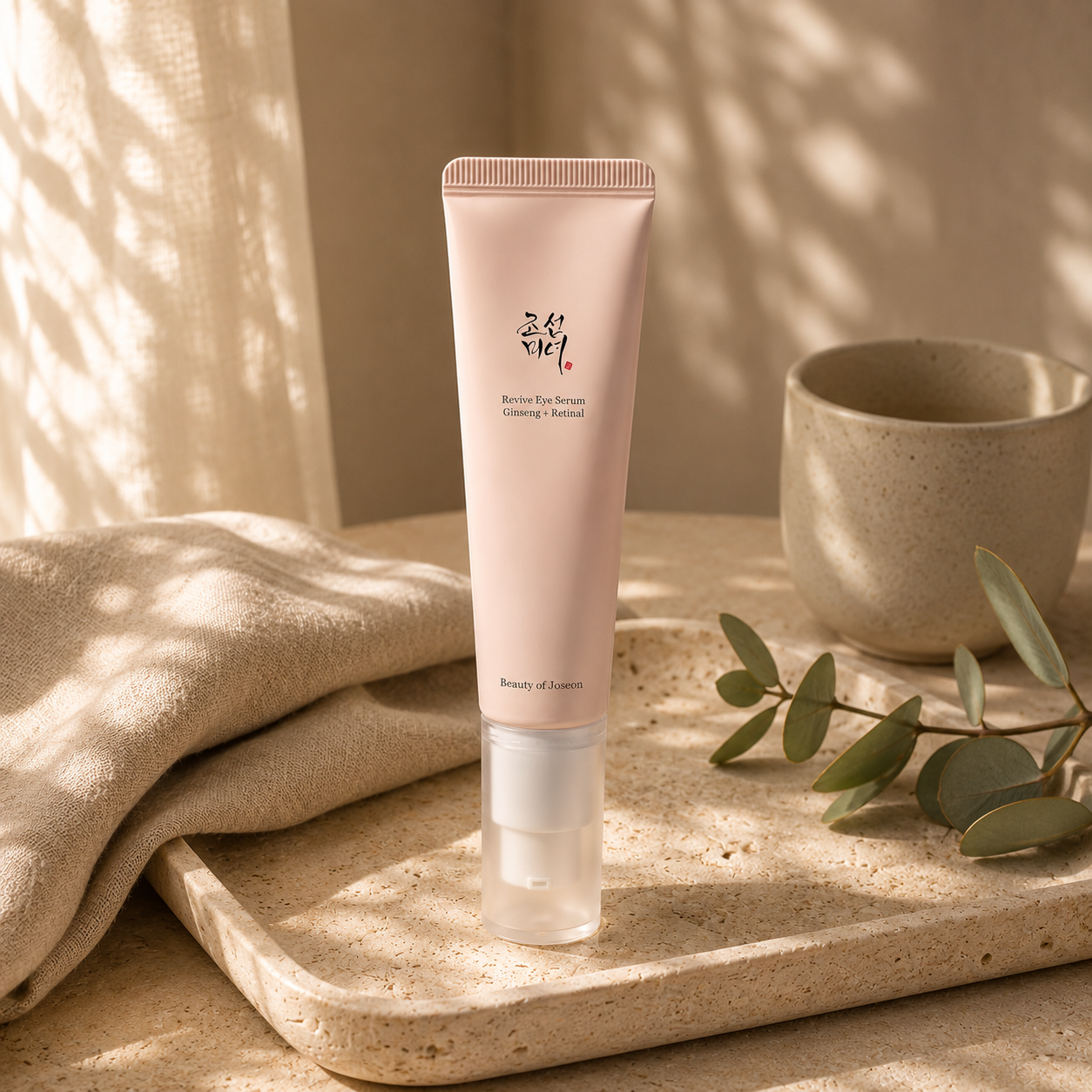Beauty of Joseon Revive Eye Serum Ginseng + Retinal displayed naturally on a warm travertine vanity with a folded beige linen cloth, ceramic cup, glass vase with eucalyptus, and soft morning sunlight, creating a premium Korean skincare lifestyle editorial scene.