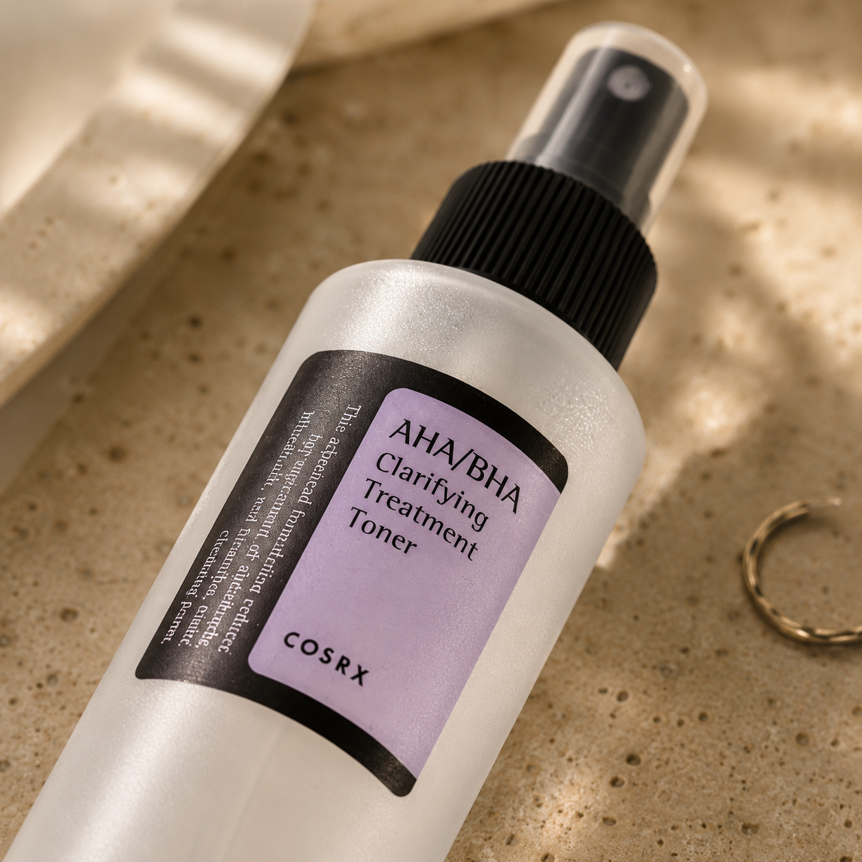 Macro close-up of the COSRX AHA/BHA Clarifying Treatment Toner bottle resting on warm beige travertine, highlighting the frosted bottle finish, fine mist spray, and label details with soft morning sunlight and a luxury Korean skincare editorial aesthetic.
