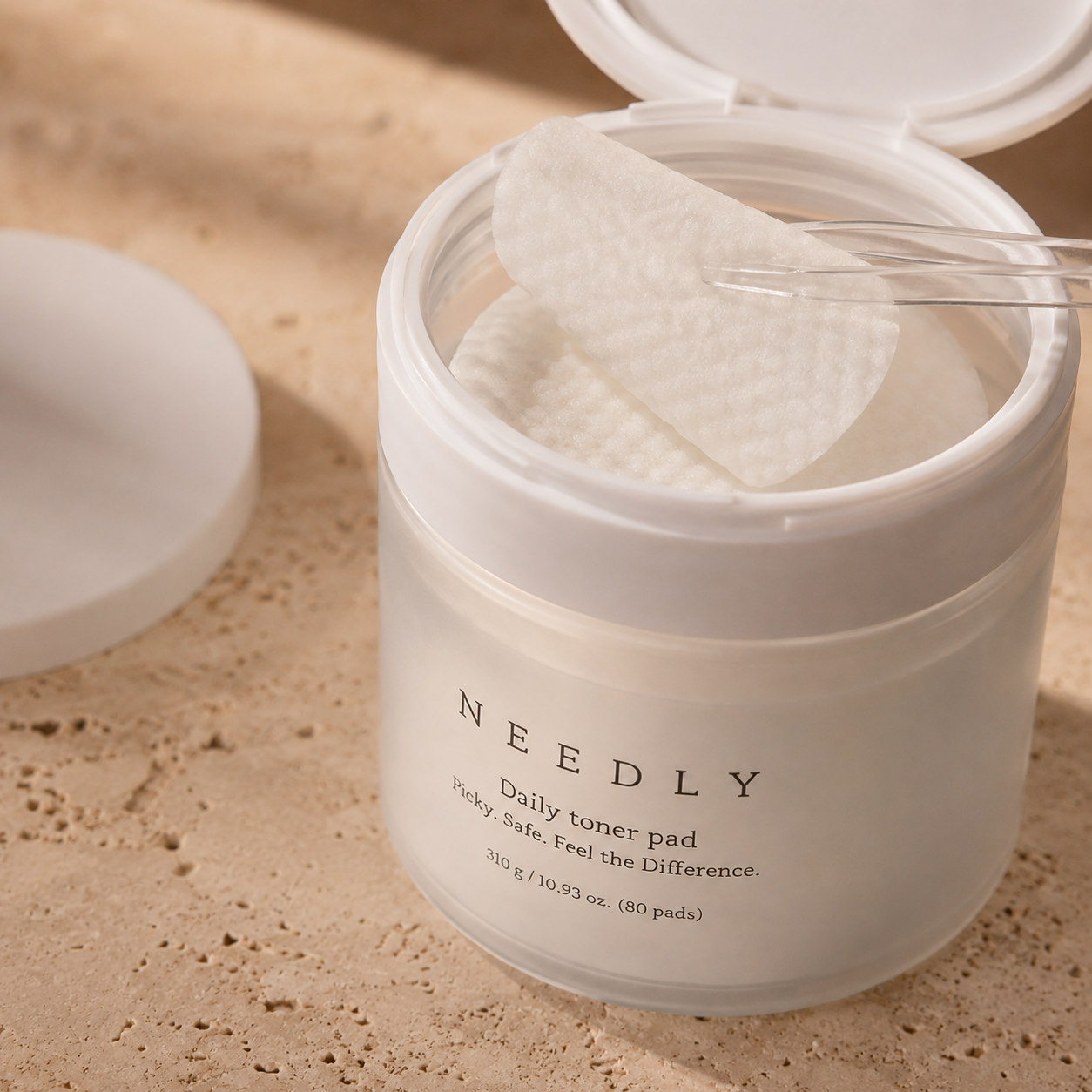 Close-up of Needly Daily Toner Pad 310g 80 pads with a textured pad lifted by clear tweezers, showing the soft embossed pad surface in warm natural light.