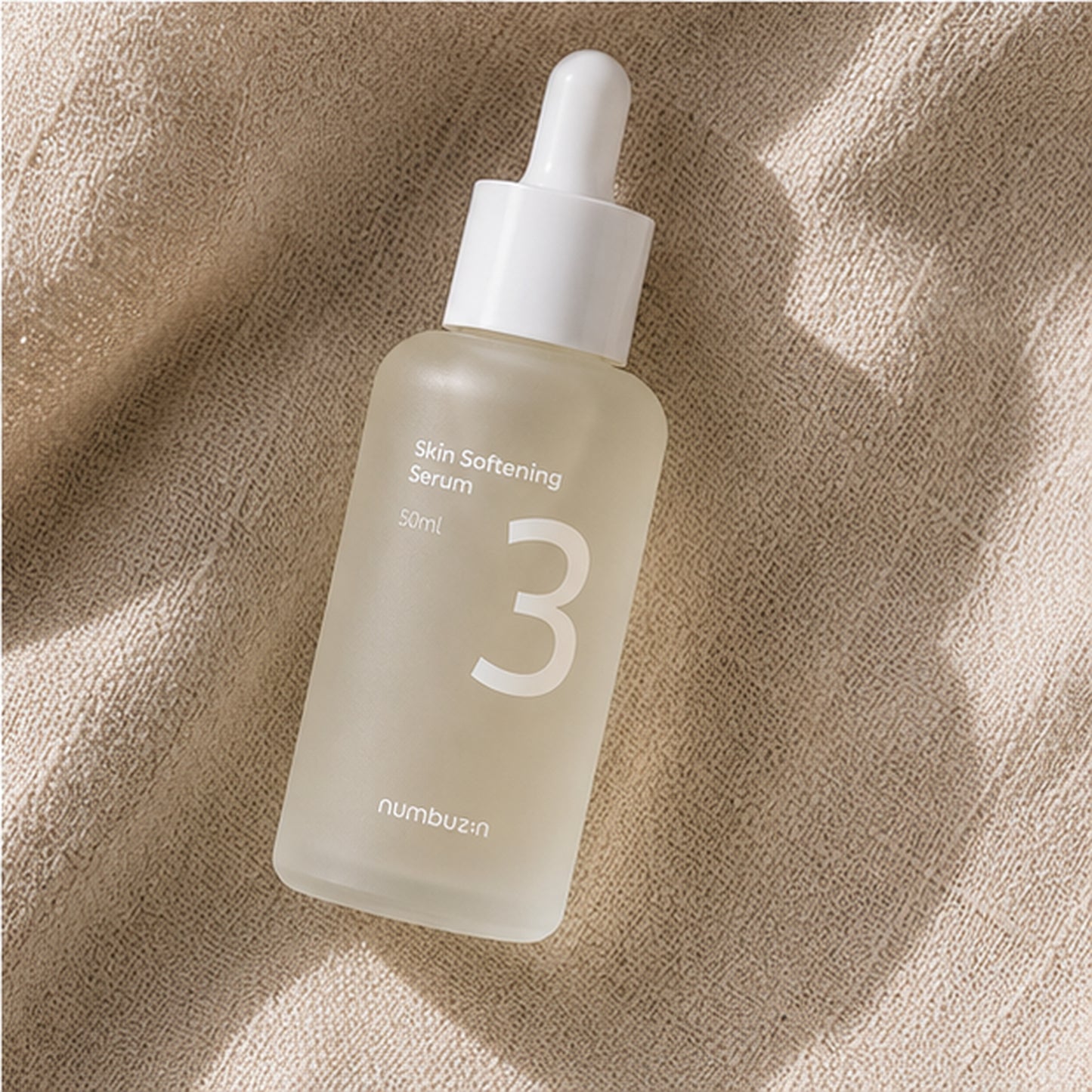Numbuzin No.3 Skin Softening Serum 50ml styled on a travertine vanity with beige linen, ceramic bowl, glass vase, and soft botanical shadows.