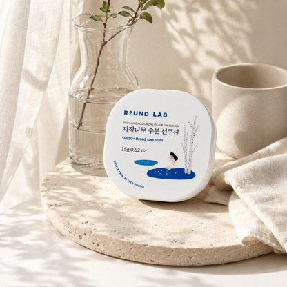 Round Lab Birch Juice Moisturizing UVlock Sun Cushion SPF50+ displayed on a travertine vanity with soft water reflections, linen, ceramic accessories, and natural morning light.
