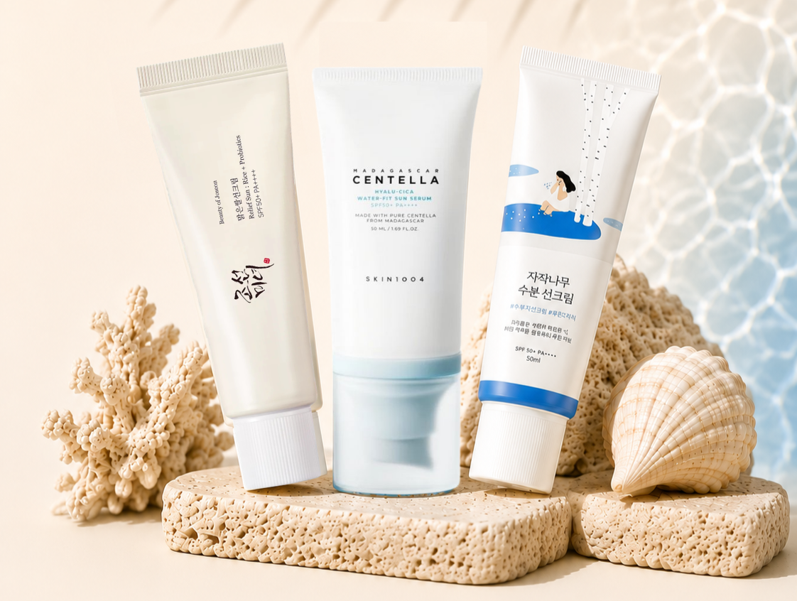 editorial summer sunscreen flat lay featuring Beauty of Joseon Relief Sun, SKIN1004 Hyalu-Cica Water-Fit Sun Serum, and ROUND LAB Birch Juice Moisturizing Sun Cream styled with coral, seashells, and soft sunlight on neutral stone pedestals.